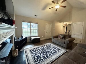 A living room with a patterned area rug, two brown couches, a TV mounted above a fireplace, a window letting in natural light, and a ceiling fan with light. A small ottoman sits near the window.