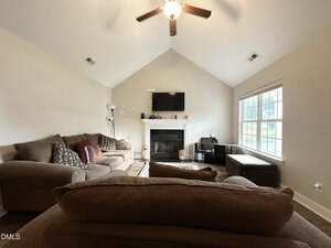 A living room with a brown sectional sofa, a fireplace topped with a TV, a lamp, decorative pillows, a large window letting in daylight, and a ceiling fan on the white vaulted ceiling.