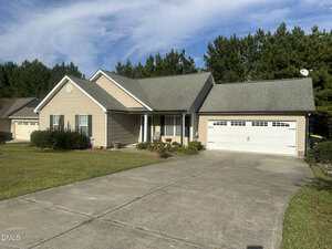 Charming single-story beige house with white trim at 109 Kinloss Way. Enjoy an attached two-car garage, concrete driveway, and a front yard filled with grass, shrubs, and a small flower bed bordered by tall pine trees that stem in the background.