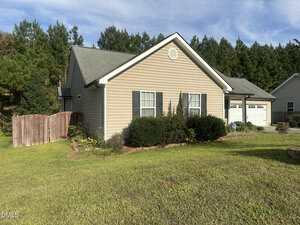 A single-story beige house with white trim, gray shingles, and two front windows. There is a garage on the right and a wooden fence on the left. The yard is grassy with some bushes by the house and trees in the background.