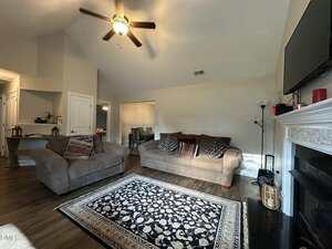 A living room with a ceiling fan, two sofas, patterned rug, and a fireplace. A TV is mounted above the fireplace. There are side tables, lamps, and decorative cushions. The dining area is visible through an open doorway.