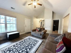A living room with a ceiling fan, two brown sofas with patterned pillows, a black and white rug, a window, and a door. The space has wood flooring and neutral-colored walls with a view into a hallway and other rooms.