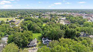 Aerial view of a suburban neighborhood with several houses surrounded by dense green trees; a small town with multiple buildings is visible in the distance under a blue sky with scattered clouds.