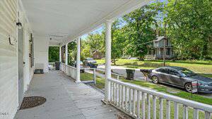 A wide front porch with white railings and columns overlooks a residential street with parked cars, trash bins, and houses surrounded by green trees.