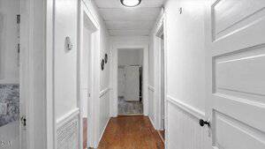 A bright hallway with white walls, white doors, and wood flooring. The hallway has three visible doorways, recessed ceiling lights, and wainscoting on the lower portion of the walls.