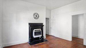 A small room with white walls, a wooden floor, and a black fireplace with a wall-mounted heater. A black and white clock hangs above the fireplace. Two white doors are visible on the right side.