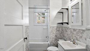 Small bathroom with a white door, bathtub with shower, window above the tub, white vanity with sink and faucet, toilet, and built-in white cabinets and shelves along the wall. Marble-patterned wall panels surround the space.