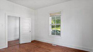 An empty room with white walls, a wooden floor, a single window with blinds, and an open white door. Sunlight comes through the window, showing part of a car and greenery outside.