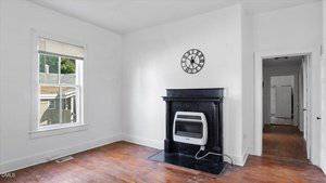 A small room with white walls and a wooden floor features a black fireplace with a wall-mounted gas heater and a round clock above it. There is a window on the left and an open doorway leading to a hallway on the right.