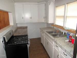 A small galley kitchen with white cabinets, marble-patterned countertops, a stainless steel gas stove, double sink, and two windows with blinds. Various cleaning supplies and items are on the countertop near the sink.