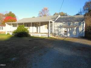 Located at 322 Aiken Avenue in Creedmoor, this single-story house features a covered front porch, white siding, and an attached garage. A gravel driveway leads to the home, bordered by a dry lawn and trees under a clear sky.