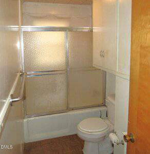 A small bathroom with a bathtub and sliding glass shower doors, a white toilet, white cabinets above the toilet, a towel bar on the left wall, and a wooden door partially opened.