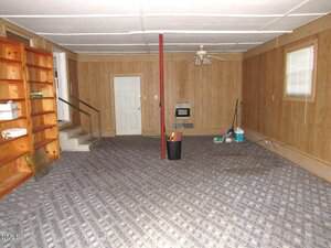 A basement room with wood-paneled walls, carpeted floor, a red support pole, built-in wooden shelves on the left, a ceiling fan, cleaning supplies in one corner, and a window on the right wall.
