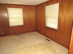 An empty room with wood-paneled walls, two windows with closed blinds, beige carpet, and visible floor vents. Electrical outlets are present on the walls. The room appears clean and undecorated.