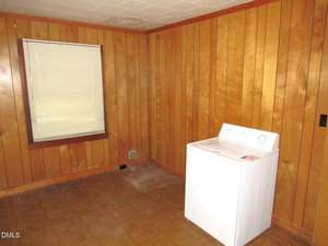 A small room with wood panel walls, a single window with closed blinds, and a white washing machine in the corner on a patterned linoleum floor. The room appears mostly empty and has some discoloration near the baseboard.