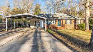 Single-story brick house with a blue front door, white trim, and attached carport. Concrete driveway extends from the street to the carport. Yard has grass, trees, and some shrubs near the house.