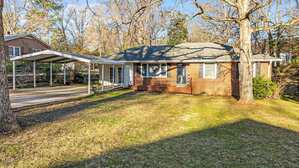 A single-story brick house with a gray shingle roof, large front windows, and an attached carport. The front yard is grassy with some patches of bare ground and bordered by mature trees.