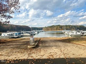 A concrete boat ramp leads down to a marina with several boats docked on the water, surrounded by trees under a partly cloudy sky. Fallen leaves are scattered along the ramp and ground.