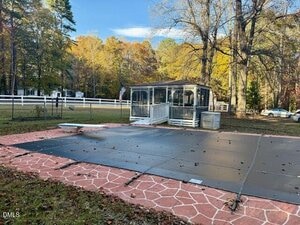 A covered in-ground pool sits in a backyard with a red and white patterned deck. Behind the pool is a white screened gazebo, surrounded by trees with autumn foliage and a white fence.