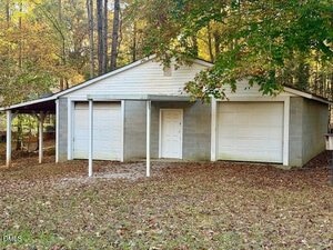 A one-story gray cinder block garage with two white roll-up doors, a white entry door in the center, three white support posts in front, and a carport on the left, surrounded by trees and fallen leaves.