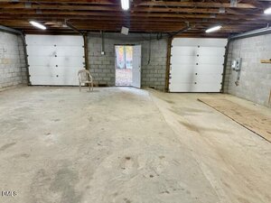 A spacious, empty garage with unfinished concrete floors, exposed ceiling beams, two closed garage doors, one open door to the outside, an electrical panel, and a single plastic chair near the wall.
