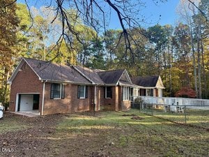 A single-story brick house with an attached garage, large front windows, and a wheelchair-accessible ramp, set on a grassy yard with trees in the background under a clear sky.