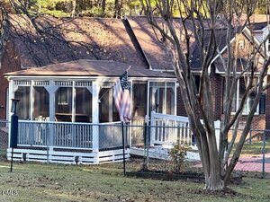 A brick house features a screened-in porch with a white railing and an American flag displayed. Leafless trees stand in the yard and the ground is covered in grass with patches of sunlight and shadow.
