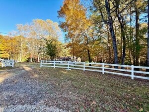 A gravel driveway curves past a white wooden fence in a yard with scattered autumn leaves and trees with yellow and green foliage. A house is partially visible in the background among the trees.