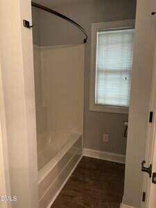 A bathroom with a white bathtub and shower combination, gray walls, a window with closed blinds, wood-look flooring, and a toilet paper holder next to the tub.