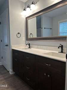 A bathroom with a double sink vanity featuring dark wooden cabinets, a white countertop, two black faucets, a large framed mirror, wall-mounted lights above, and a towel ring next to a white door.