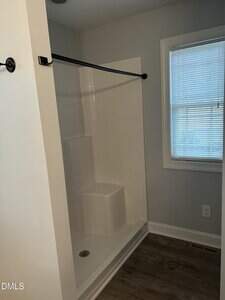 A small bathroom with a walk-in shower featuring a built-in seat, white tile walls, a black shower curtain rod, a window with blinds, and wood-look flooring.