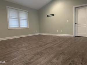 An empty room with light green walls, a white ceiling, wood-style flooring, a window with blinds, electrical outlets, an air vent, and a closed white door.