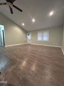 A spacious, empty room with light green walls, wood flooring, recessed ceiling lights, a ceiling fan, a window with closed blinds, and a door partially covered by a sheer curtain blowing inward.