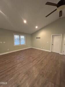 A spacious, empty room with light green walls, wood flooring, a white door, a window with blinds, and a ceiling fan. The room has recessed lighting and a vaulted ceiling.