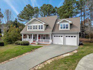 Two-story house with gray siding, white trim, and a covered front porch. There are two garage doors on the right and a wide concrete driveway. The yard is grassy with some bushes and surrounded by trees in the background.