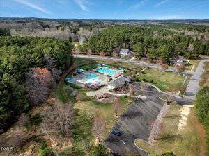 Aerial view of a community area with a swimming pool, lounge chairs, umbrellas, a clubhouse building, a parking lot, a small playground, and surrounding trees and houses. Rural landscape extends in the background.