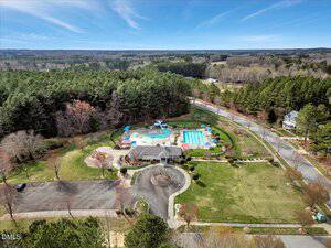 Aerial view of a community recreation area with two outdoor swimming pools, lounge chairs, shade umbrellas, a clubhouse building, surrounding lawn, trees, and a nearby road, bordered by a forest.