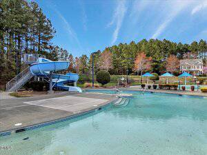 Outdoor swimming pool with a curved blue water slide on the left, several steps leading into the shallow end, and blue umbrellas shading tables in the background. Trees and a large house are visible behind the pool area.