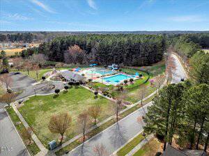 Aerial view of a community pool and playground area surrounded by trees and grass, with several roads and parked cars nearby on a clear day.