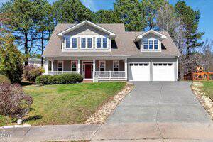 A two-story suburban house with gray siding, white trim, a large front porch, two dormer windows, and a double garage. The driveway is concrete and the front yard has grass and low shrubs. Tall trees are in the background.