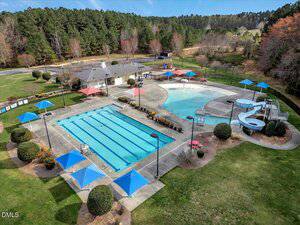 A community pool area with a rectangular lap pool, a second pool with a water slide, blue sunshades, seating areas, a small building, and surrounding grass and trees. The setting is outdoors on a clear day.