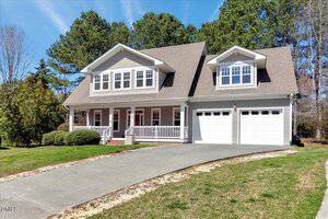 A two-story house with light gray siding, a large front porch, two dormer windows, and a double garage sits on a curved driveway with grass and trees in the background under a blue sky.