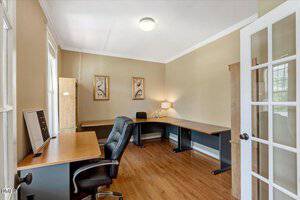 Home office with hardwood floors, beige walls, a black office chair, two wooden desks, a computer monitor, a lamp, framed art on the wall, and a glass-paned door letting in natural light from nearby windows.