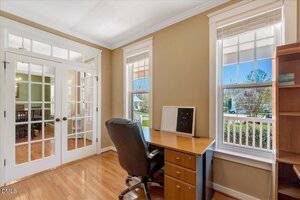A home office with a wooden desk, black office chair, and shelving, set by large windows with white trim letting in natural light. French doors open to an adjacent room with a dining table and chairs.