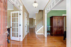 Bright entryway with hardwood floors, beige walls, white trim, French doors to the left, a staircase straight ahead, and a room with green walls and a wooden armoire to the right.