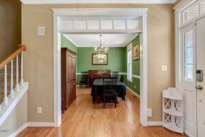 A bright dining room with green walls, wood floors, a dark rectangular table with black chairs, and a chandelier, viewed from an entryway near stairs and a white front door.