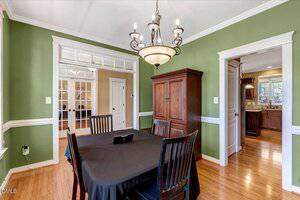 A dining room with green walls, white trim, hardwood floors, a dark wooden table with four chairs, a wooden armoire, and a chandelier. The room connects to a kitchen and an office or living area with glass-paned doors.
