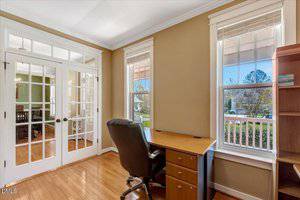 A bright home office with light brown walls, wood floors, a black office chair, and a wooden desk near two large windows. Glass French doors lead to another room with similar flooring.