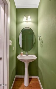 A small green bathroom with a pedestal sink, oval mirror, wall-mounted light fixture, towel ring, and a soap dispenser on the sink. The floor is wood, and the walls are painted green.