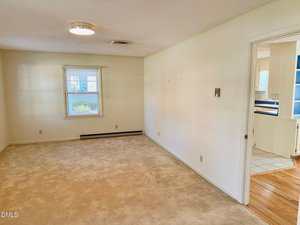 A mostly empty room with beige carpet, off-white walls, a window with blinds, a baseboard heater, and a ceiling light. An open doorway leads to a kitchen with tile flooring and visible cabinets.